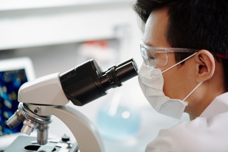 Scientist wearing face mask and goggles examining samples through a microscope in a lab