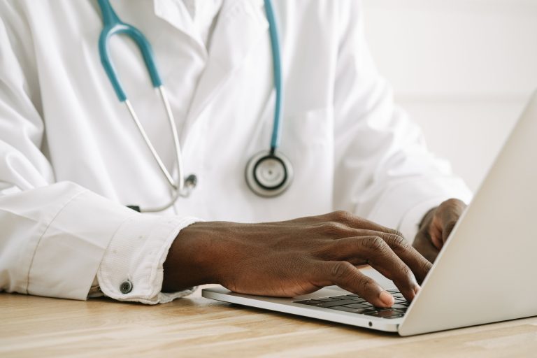 A doctor types on a laptop beside a stethoscope