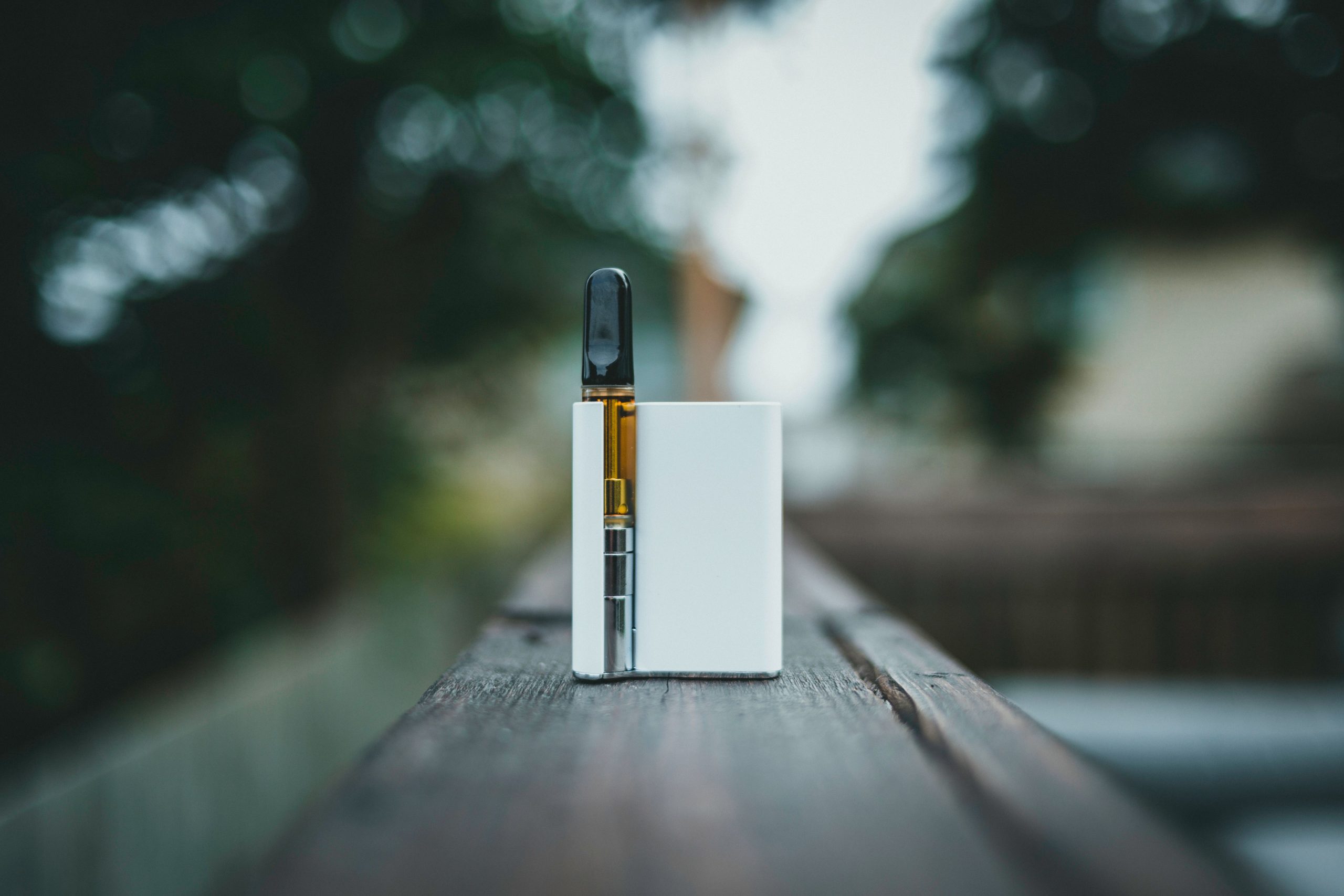 A white electronic vape device with a transparent cartridge rests on a wooden rail outdoors